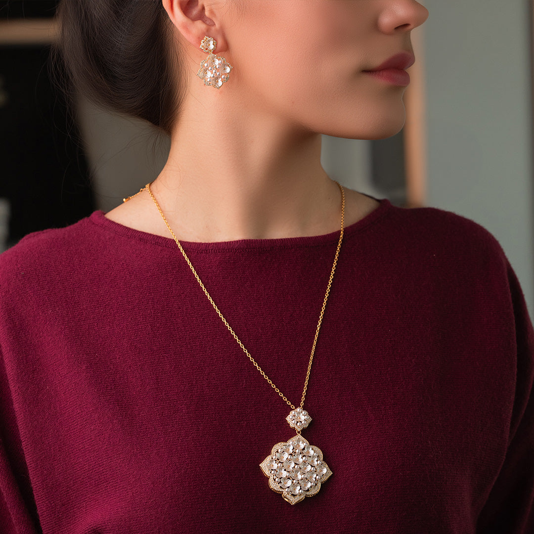 A person wearing a gold-plated necklace and earrings set with zircon stones and mother pearl, paired with a red top.