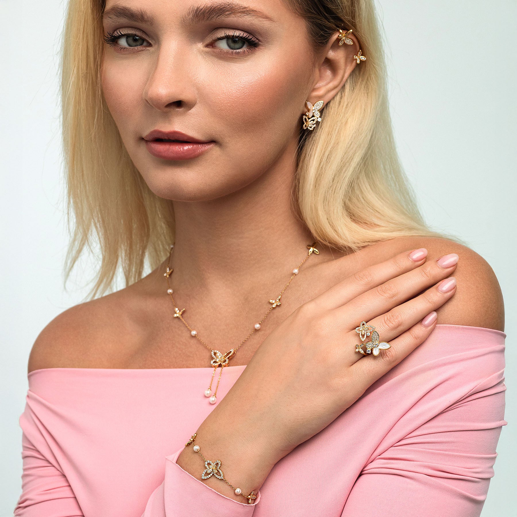 Woman wearing gold jewelry including necklace, earrings, bracelet, and ring on a light gray background