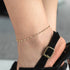 A person wearing a gold-colored beaded anklet with a metallic clasp, on a foot protruding from a black shoe.