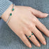 Hand wearing a silver bracelet and ring with green gemstones on a gray textured fabric background