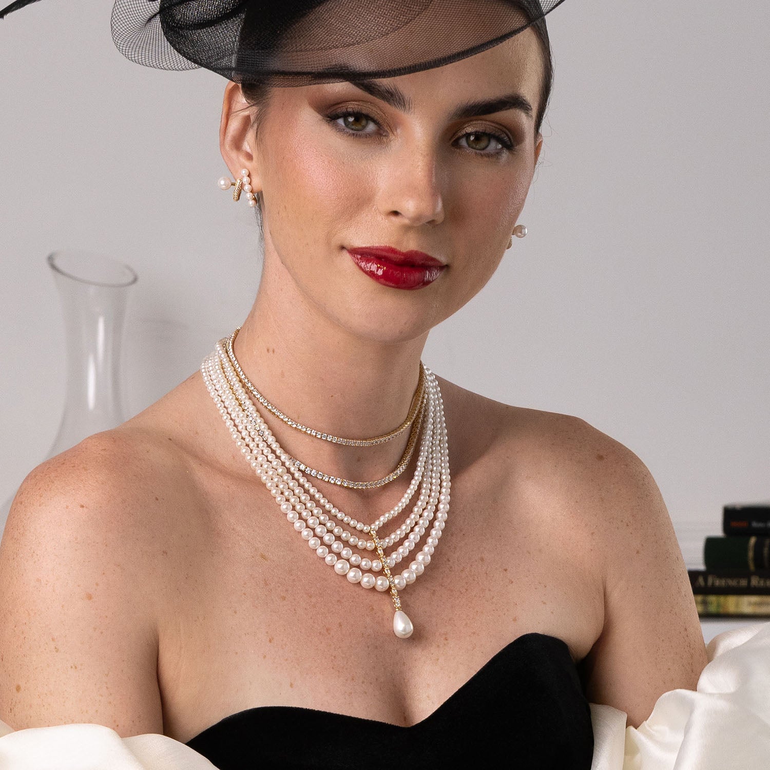 Woman wearing a pearl necklace with a black hat and red lipstick against a neutral background