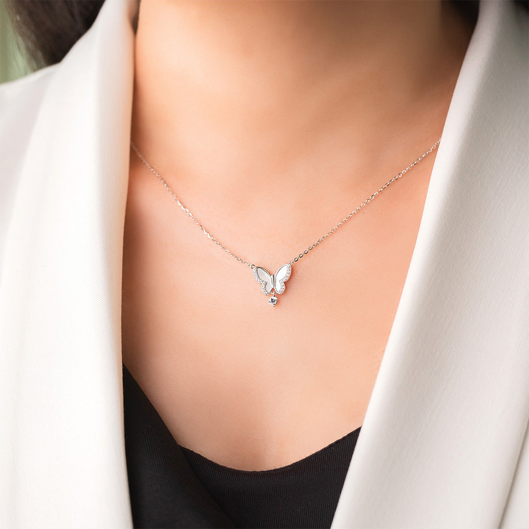 A person wearing a white blazer and a black top is showcasing a sterling silver necklace with a gold plating, featuring a butterfly pendant and small zircon stones.