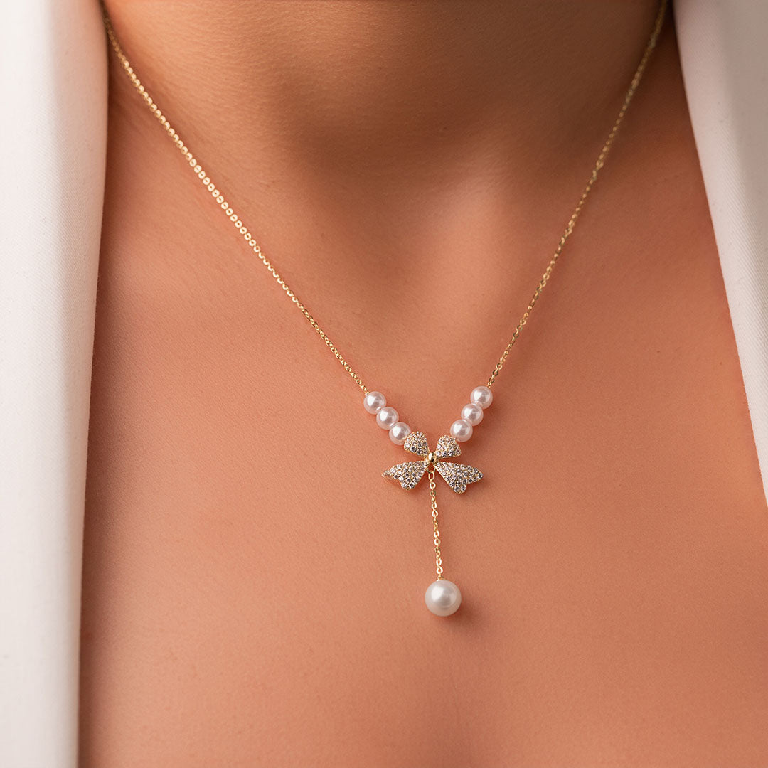 A gold-plated sterling silver necklace with a bow-shaped pendant featuring zircon stones and a faux pearl, displayed on a mannequin wearing a white coat.